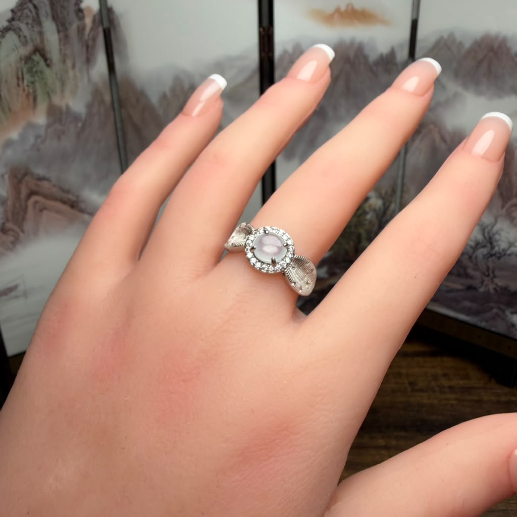 A silver ring with a round pale pink cabochon center stone and a halo of small clear stones, worn on a person's finger.