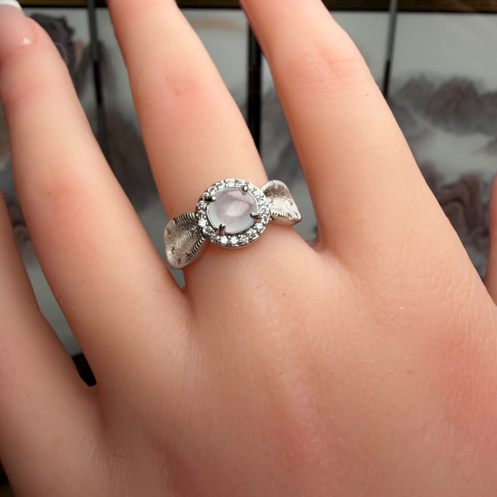 A silver ring with a round pale pink cabochon center stone and a halo of small clear stones, worn on a person's finger.