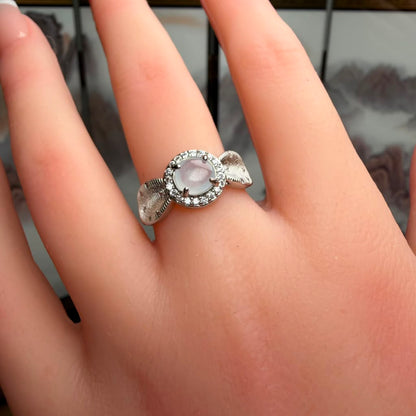 A silver ring with a round pale pink cabochon center stone and a halo of small clear stones, worn on a person's finger.