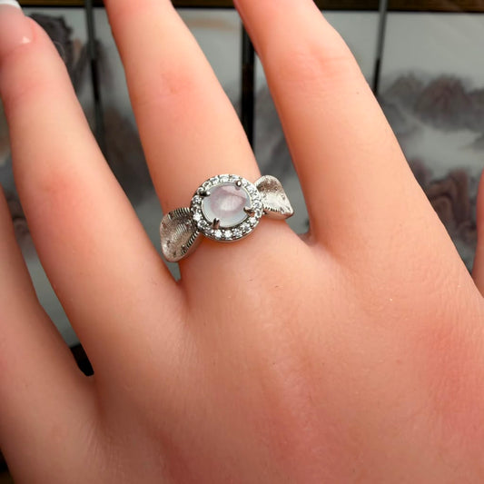 A silver ring with a round pale pink cabochon center stone and a halo of small clear stones, worn on a person's finger.