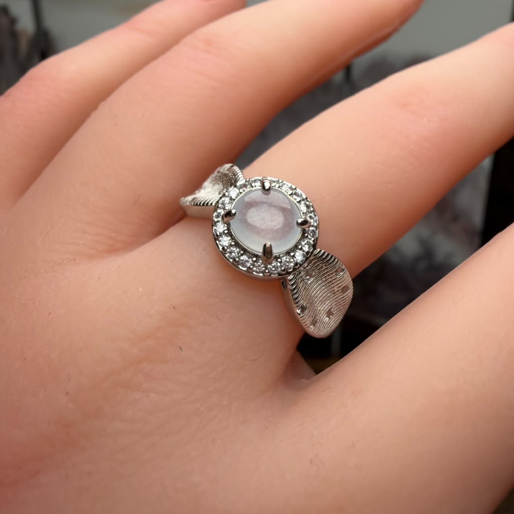 A silver ring with a round pale pink cabochon center stone and a halo of small clear stones, worn on a person's finger.