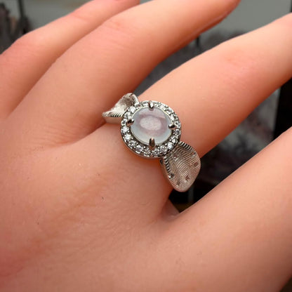 A silver ring with a round pale pink cabochon center stone and a halo of small clear stones, worn on a person's finger.